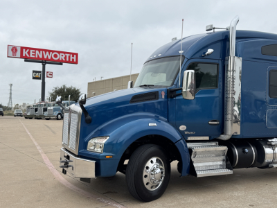 Kenworth T880 truck in blue, front-left view, with chrome exhaust stack. Visible text includes KENWORTH sign in background, vehicle features, and dealership lot.