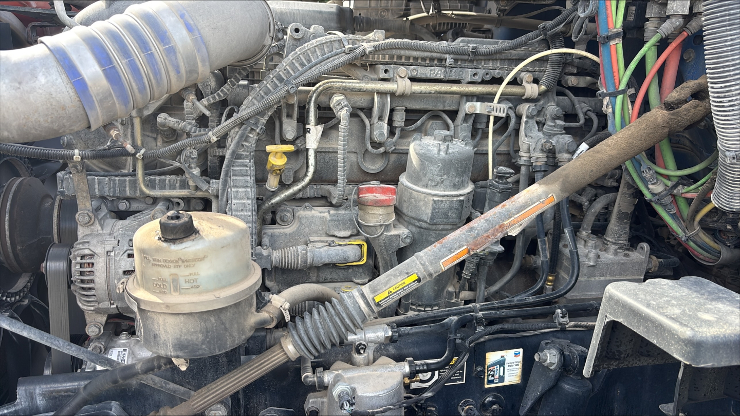 Kenworth T680 engine bay showing diesel fuel lines, reservoir caps, hoses, and electrical wiring harnesses visible under hood.