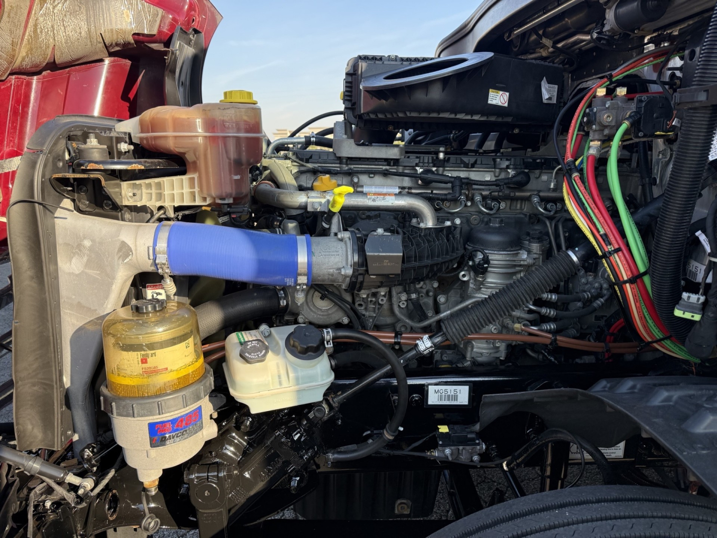 Engine bay of a 2021 Freightliner Cascadia126 with visible blue silicone hose, coolant reservoir, oil filter, and wiring harnesses; side view of the engine compartment.