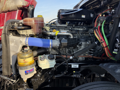 Engine bay of a 2021 Freightliner Cascadia126 with visible blue silicone hose, coolant reservoir, oil filter, and wiring harnesses; side view of the engine compartment.
