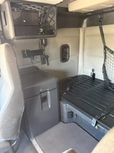 Truck sleeper interior showing storage cabinet, mounted monitor bracket, wall panel with small oval access, and a bed platform with ribbed surface.