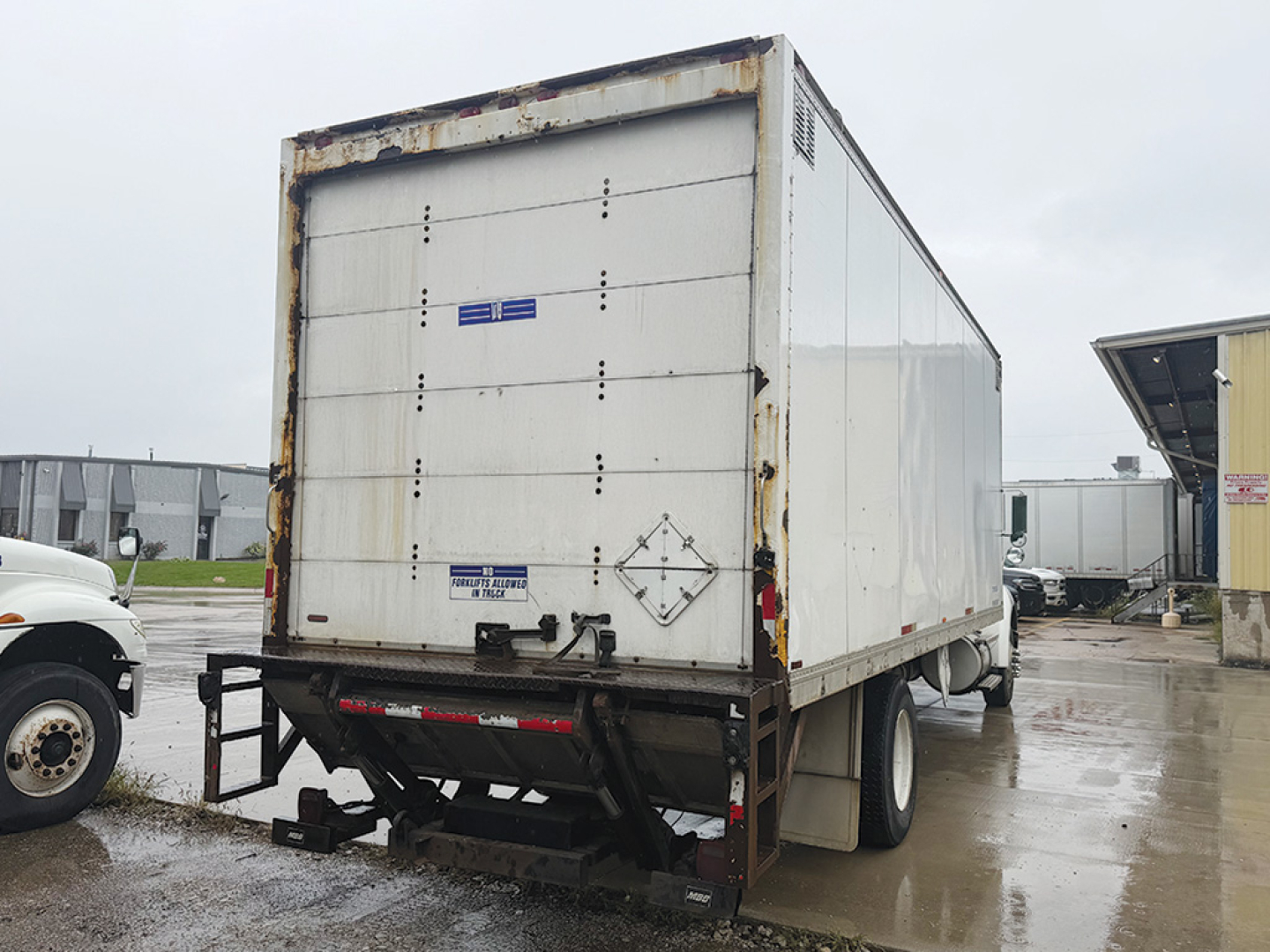 Rear view of a white Kenworth T-300 box truck trailer with rusty edges and a black landing gear, showing closed rear doors and a small blue sticker.