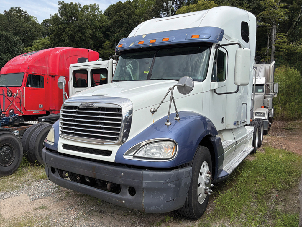 Truck: white Freightliner CL120 Columbia day cab tractor with blue lower hood and bumper, front grille, side mirrors; other trucks in background; outdoor lot.