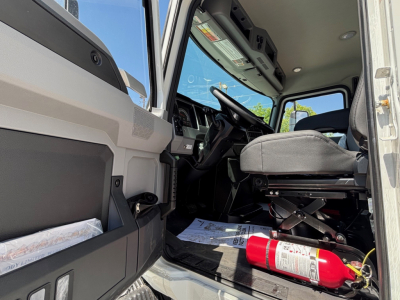 Cab interior of a new 2026 Mack MD6 truck showing driver seat, steering wheel, and dashboard from open door; light gray trim and floor mats visible.