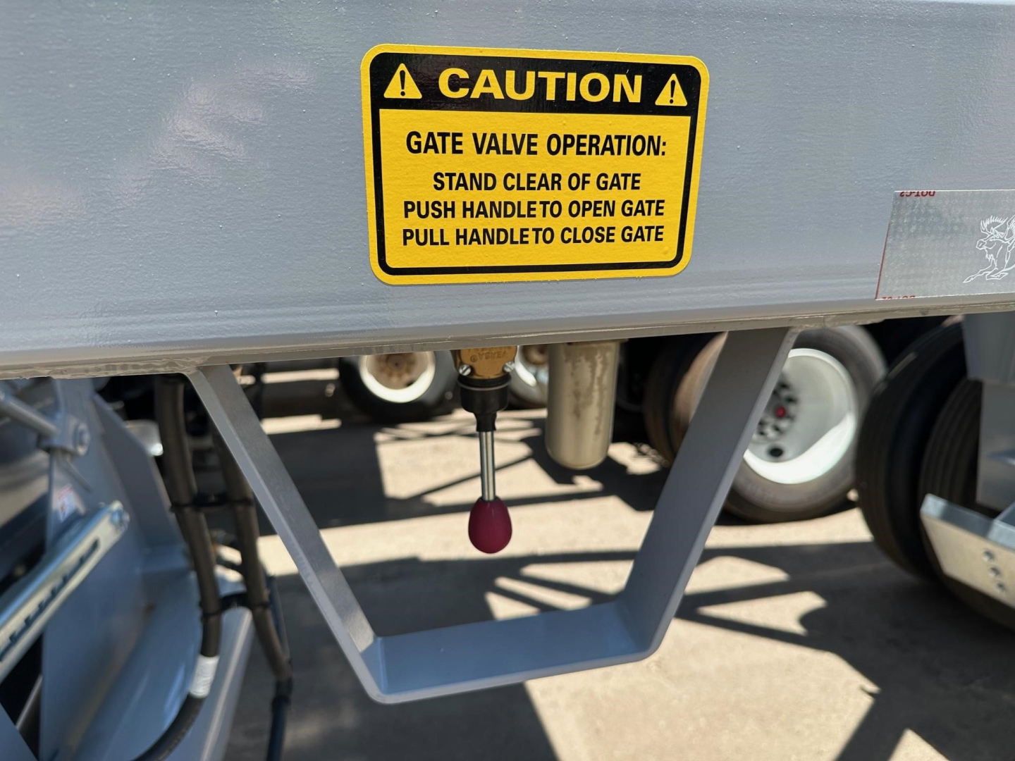 Caution label on gate valve of bottom-dump trailer with stand-off gate and red pull handle; yellow warning sticker reading CAUTION GATE VALVE OPERATION STAND CLEAR OF GATE PUSH HANDLE TO OPEN GATE PULL HANDLE TO CLOSE GATE.