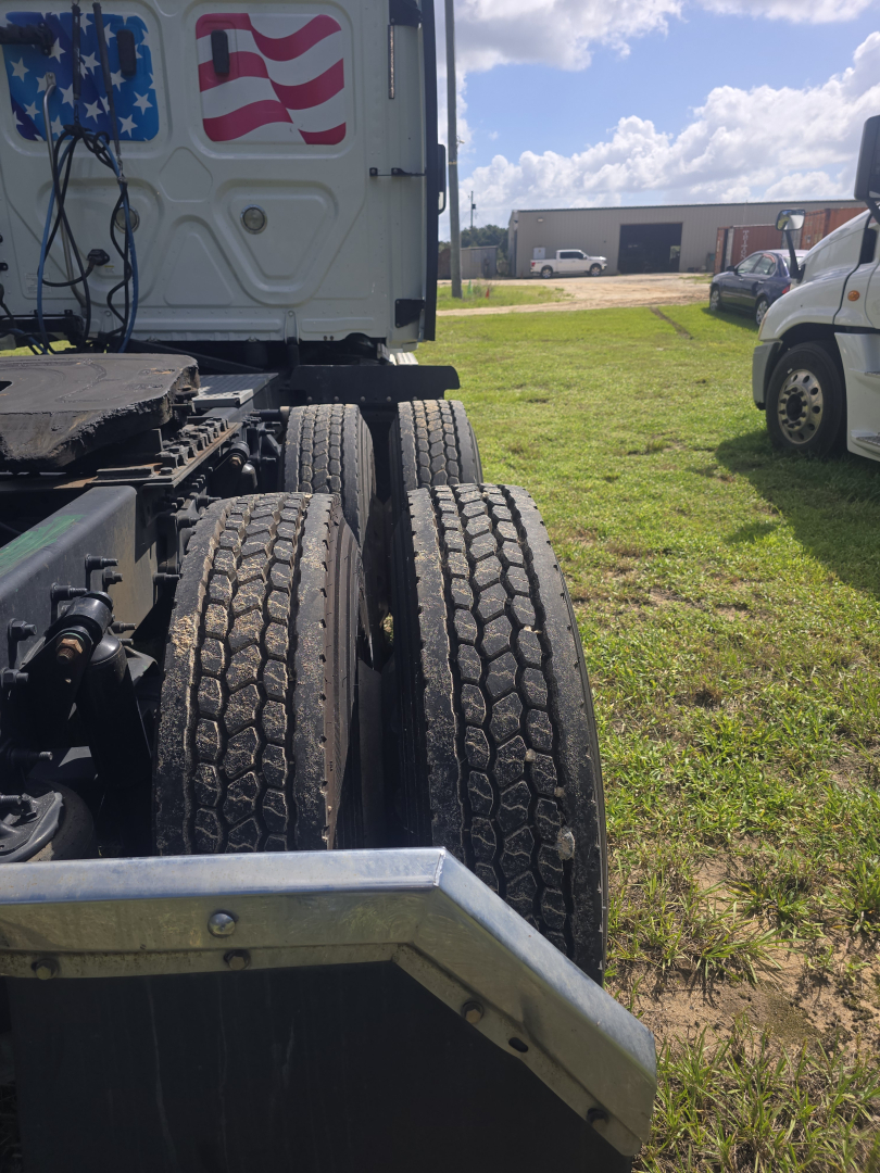 Rear view of a Freightliner Cascadia truck showing dual rear axles with large tires; partial American flag graphic on the cab; warehouse and other vehicles in background.