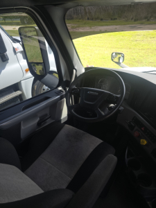 Cab interior of a Freightliner Cascadia truck showing steering wheel, driver seat, and dashboard in view.