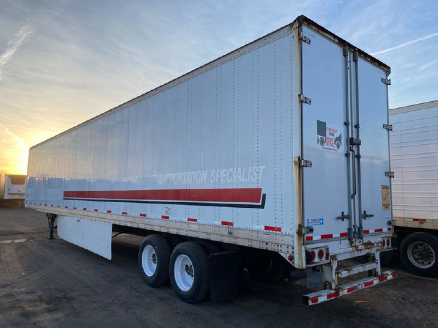 trailer body with double rear doors showing Stoughton logo and rear bumper, side lettering reading 'TRANSPORTATION SPECIALIST' and red stripe, tandem axle trailer