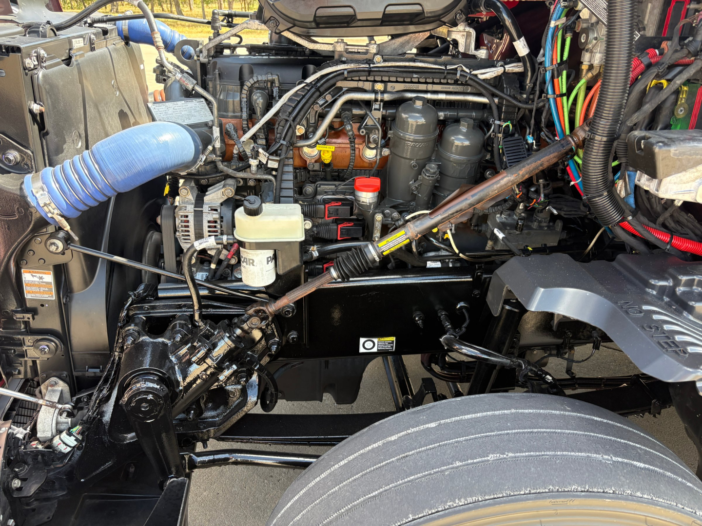 Kenworth T680 engine bay view showing fluid reservoirs, hoses, and components; visible components include coolant reservoir, battery area, air lines, and wiring harnesses.