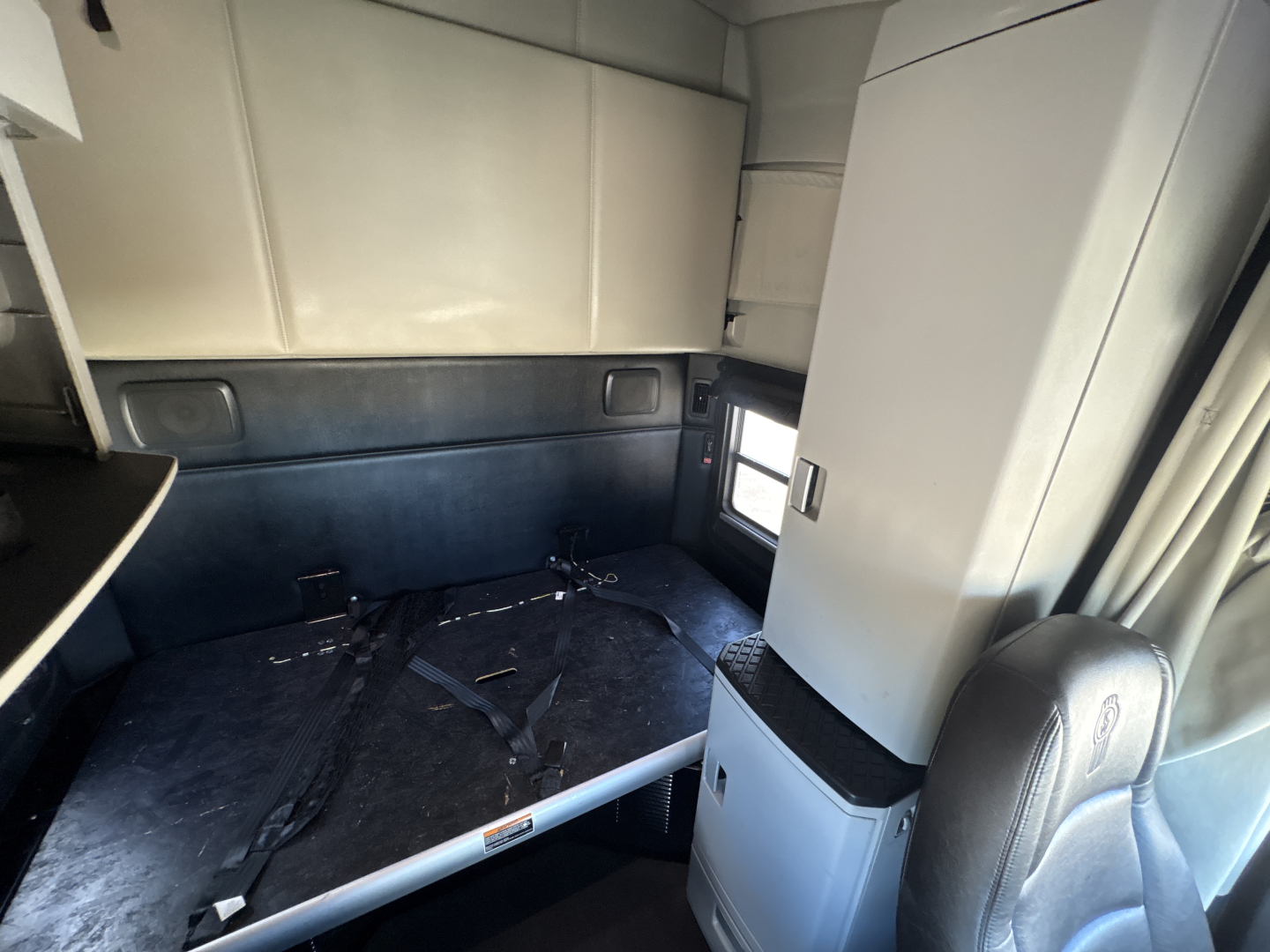 Interior of a 2023 Kenworth T680 sleeper cab showing cargo area with seat and storage compartments, black flooring, seat belt buckles, light gray cabinets, and a window