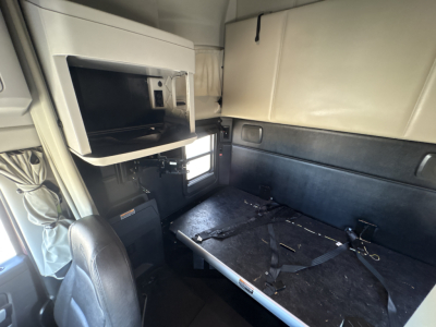 Kenworth T680 sleeper cab interior showing storage locker, overhead shelf, worktable with seatbelt and wiring, beige headliner, and dark-blue side panels.