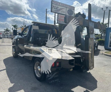 Tow truck with a mounted crane/winch in service showing the boom and winch housing on the right side; cab visible with side mirror, vehicle number placards faint on door, dealership signage in background.