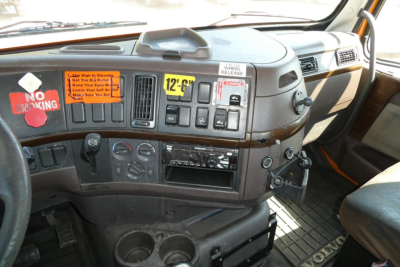 Dashboard of a Volvo truck cab with warning stickers and various gauges; visible labels include a bright orange ‘NO SMOKING’ sticker and several small placards; plastic dashboard clutter and cup holders present.
