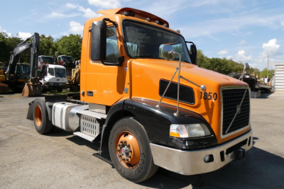 Volvo VNM42T conventional daycab truck with orange cab and black front fenders, showing wheel and bumper.