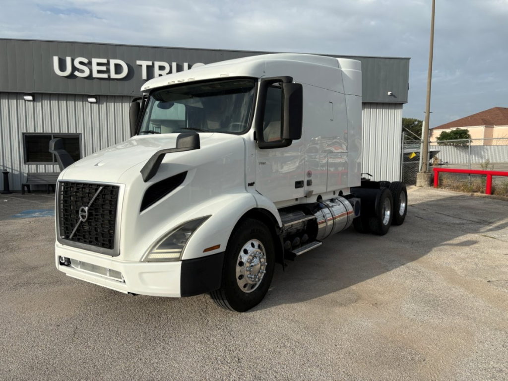 Volvo VNR64T640 tractor-trailer in white, front-left view, with sleeper cab and dual rear axles, parked in a lot near a USED TRUCKS building.