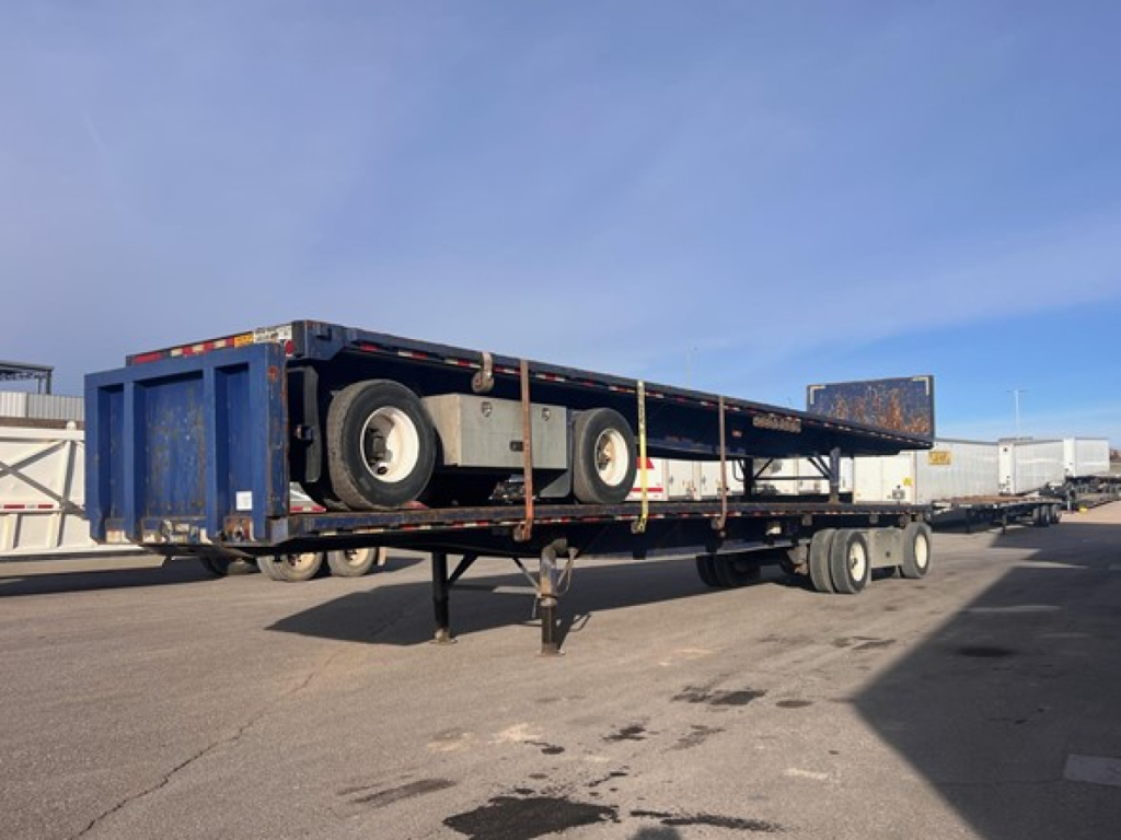 Flatbed trailer with dual tandem axles and blue sides, parked in an outdoor lot. Side ramps folded, trailer on a lowboy with support legs down.