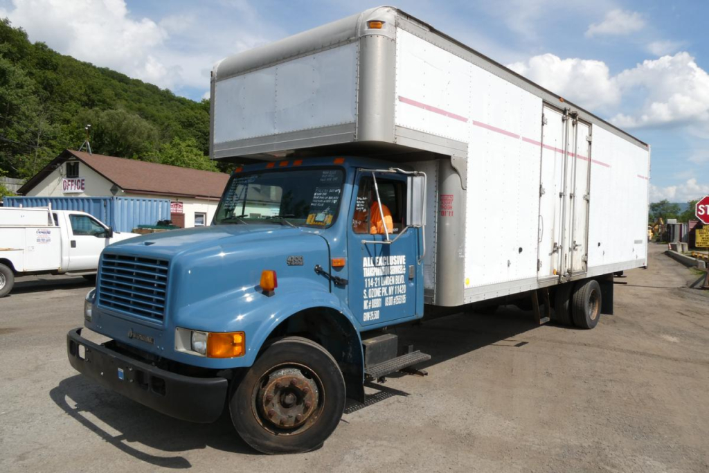Box truck with blue cab and white cargo box, front left view. Visible text on door and a raised cargo box on International 4700.