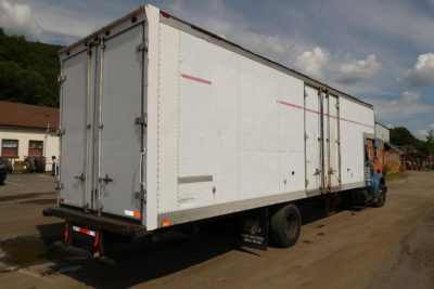 Box truck with white cargo box and blue cab seen from rear-right angle, shows long rectangular box with light pink reflective tape, metal hinges, cargo doors, and rear bumper.
