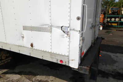 Truck trailer rear corner showing white box body with metal diamond plate step, hinge, latch, and bumper, on a trailer bed with site background.