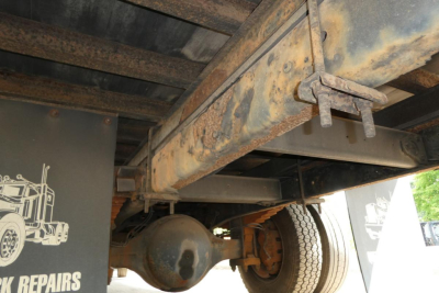 Heavy-duty truck chassis underside with leaf springs and axle housing visible, rust-toned metal frame and rear wheel in view.
