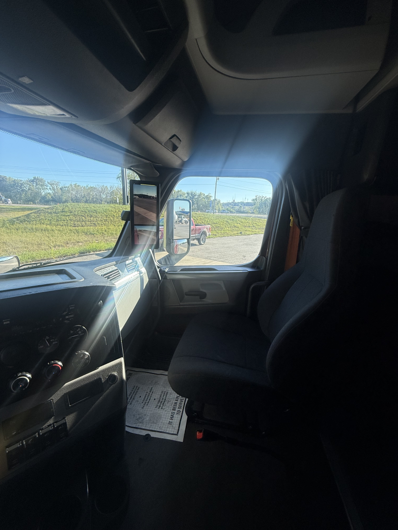 Truck cabin interior with dashboard controls visible on left and passenger seat on right; windshield shows side mirror and outside scenery.