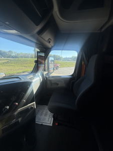 Truck cabin interior with dashboard controls visible on left and passenger seat on right; windshield shows side mirror and outside scenery.