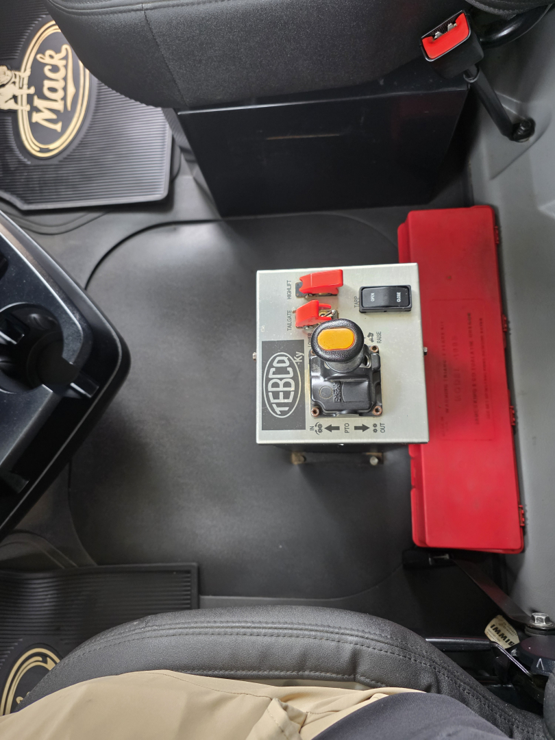 Mack Granite truck interior showing a control panel with red levers and a yellow circular knob mounted on a metal box, near a red storage container.