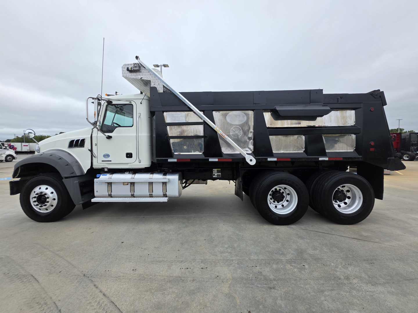 Dump truck side view showing Mack GRANITE dump body with black container and white cab. Visible text on side includes no specific legible labels; general components: front grille, exhaust stack, fuel tanks under cab, step and ladder.