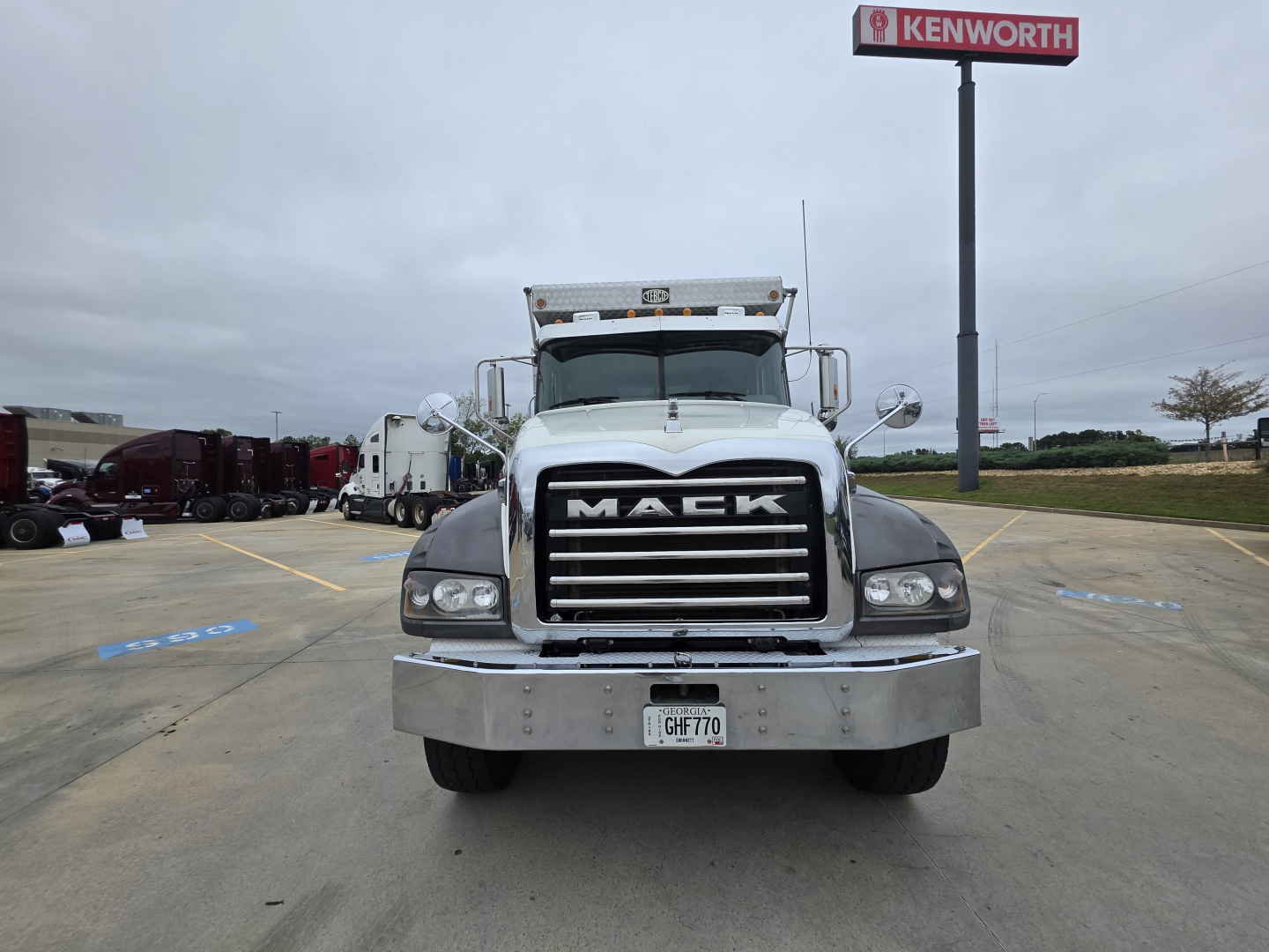 Mack Granite front view showing bold grille and bumper with Mack emblem, parking lot background with Kenworth sign, California plates GHFT70.