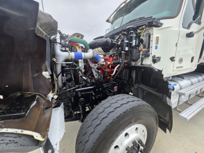 Engine bay of a 2021 Mack Granite dump truck showing engine components, hoses, radiator area, and chassis near front wheel.