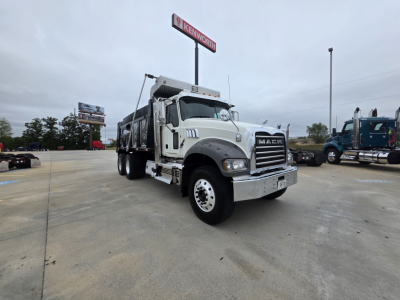 Mack Granite dump truck in a dealership lot showing front grille with Mack emblem and large chrome bumper, white cab, dump bed, and Kenworth sign in background.