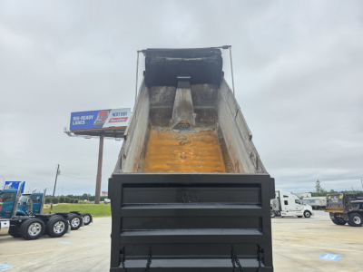 Dump truck bed with raised orange-tinted bed liner and open tailgate, Mack GRANITE likely on site, multiple trucks in background.