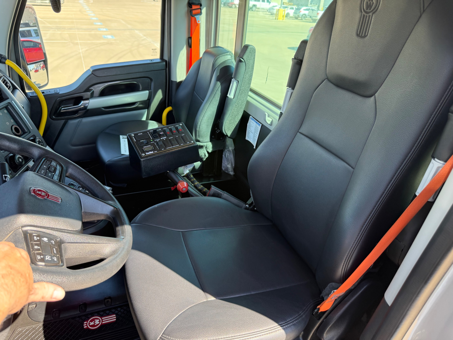 Kenworth T880 mixer truck interior showing black fabric seats, yellow steering wheel accent, dashboard with control panel, and orange seat belt. Driver seat and passenger area visible.