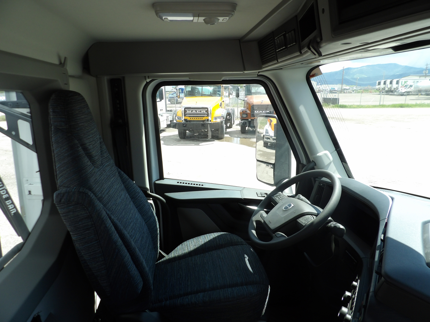 Volvo truck cab interior view showing steering wheel, driver seat, dashboard, and skylight; other trucks visible through the windshield.