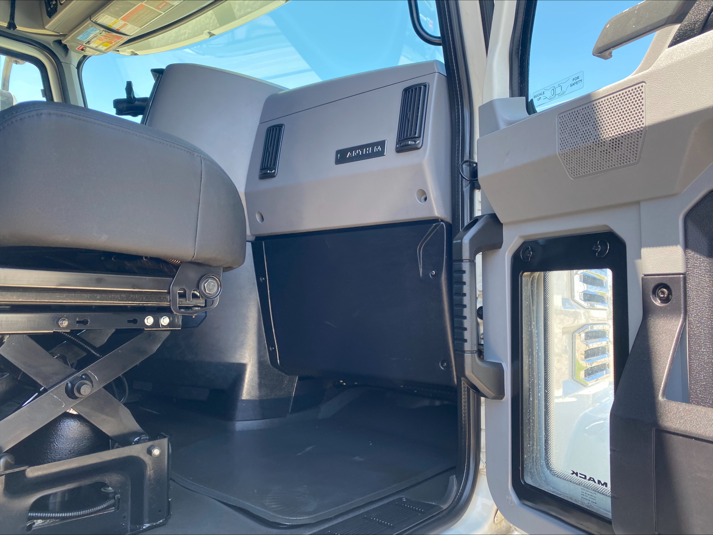 Truck cab interior showing driver's seat and dashboard with 'APX/EM' badge, door panel, and window reflection, 2019 Mack AN64T70 cab area.