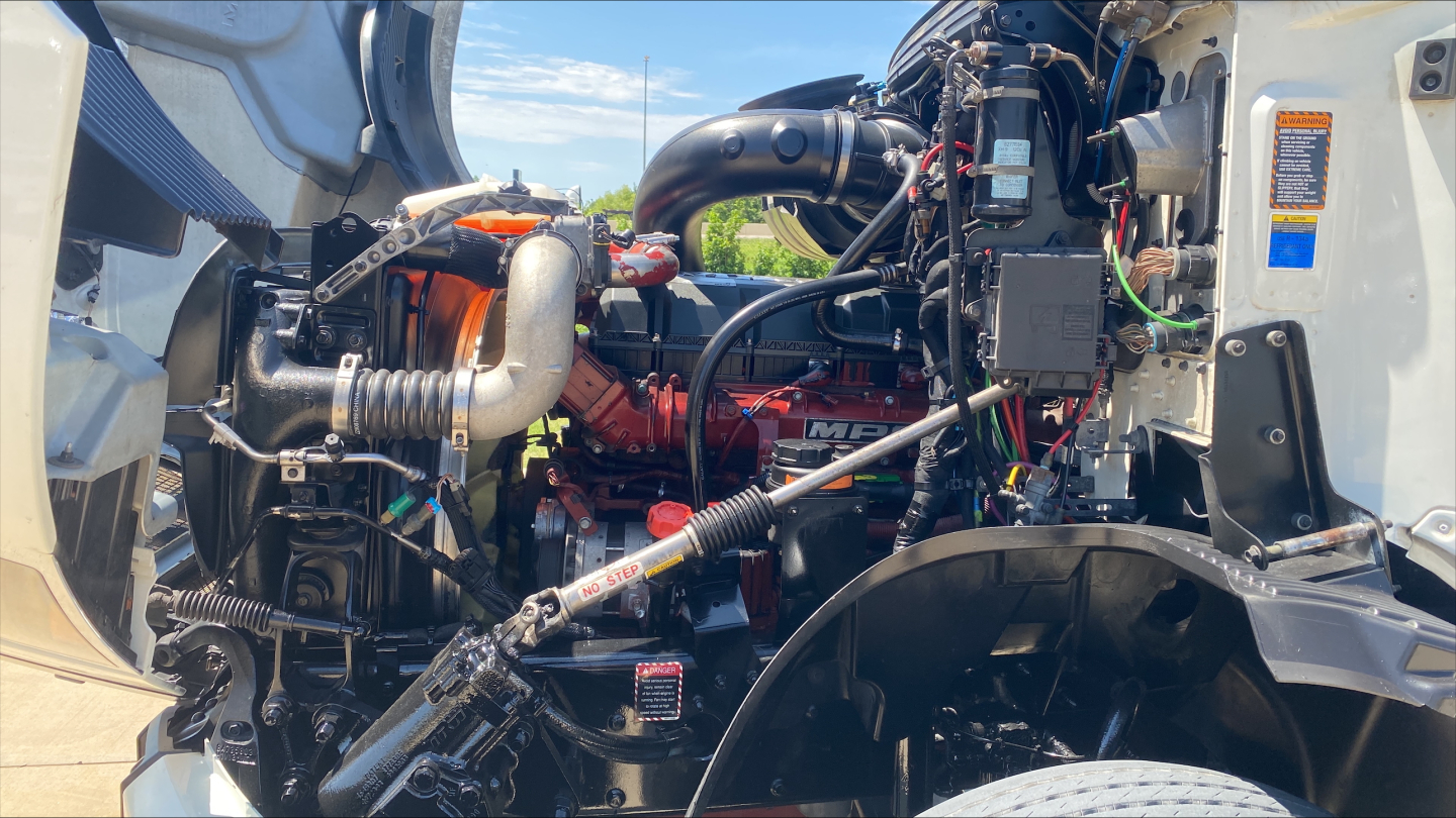 Mack AN64T70 engine bay showing diesel engine with labeled hoses, wires, and components in truck front, visible air intake, turbo piping, and electrical harnesses.