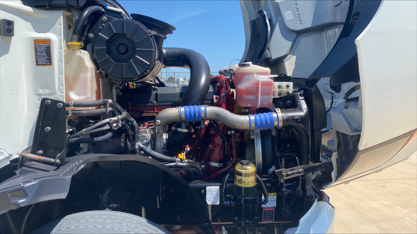 Engine bay of a 2019 Mack AN64T70 conventional sleeper showing radiator hoses, coolant reservoir, intake piping, and turbo components.