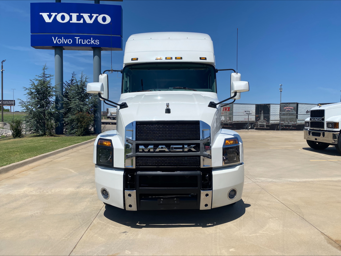 Mack AN64T70 conventional sleeper truck front view with large grille, white cab, bumper guards, and Volvo Trucks sign in background.