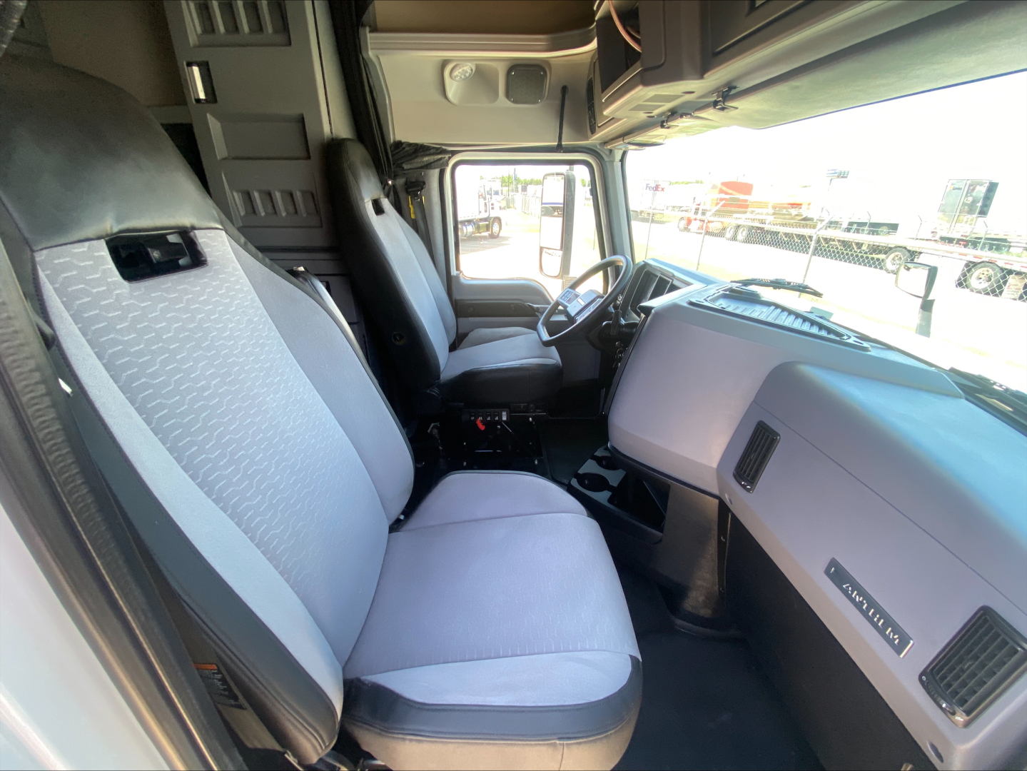 Cab interior of a 2019 Mack AN64T70 truck showing front seats, steering wheel, and dashboard. Visible text includes Mack branding on the glove area; light gray seats and dark gray floor.