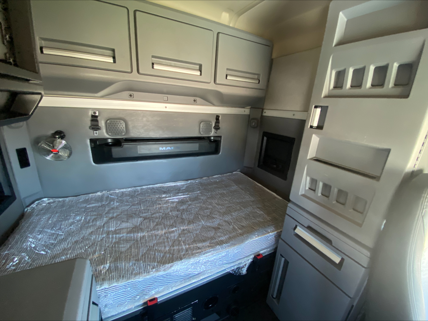 Truck sleeper cab interior showing a bed with protective cover, overhead cabinets, and storage compartments in a Mack AN64T70.