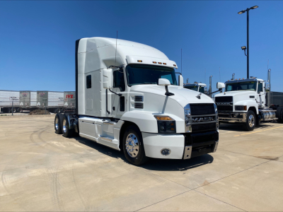 Conventional sleeper tractor showing Mack AN64T70 with white cab and cargo trailer racks in background.