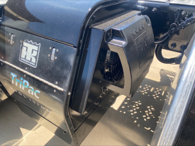 Truck engine bay area showing a compact air intake or coolant reservoir with black housing, wiring, and Tripac label on side panel of a Mack AN64T70 sleeper.