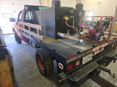 Flatbed truck with industrial auxiliary equipment mounted on the bed, rear view showing pump system, hoses, and metal diamond plate surface.