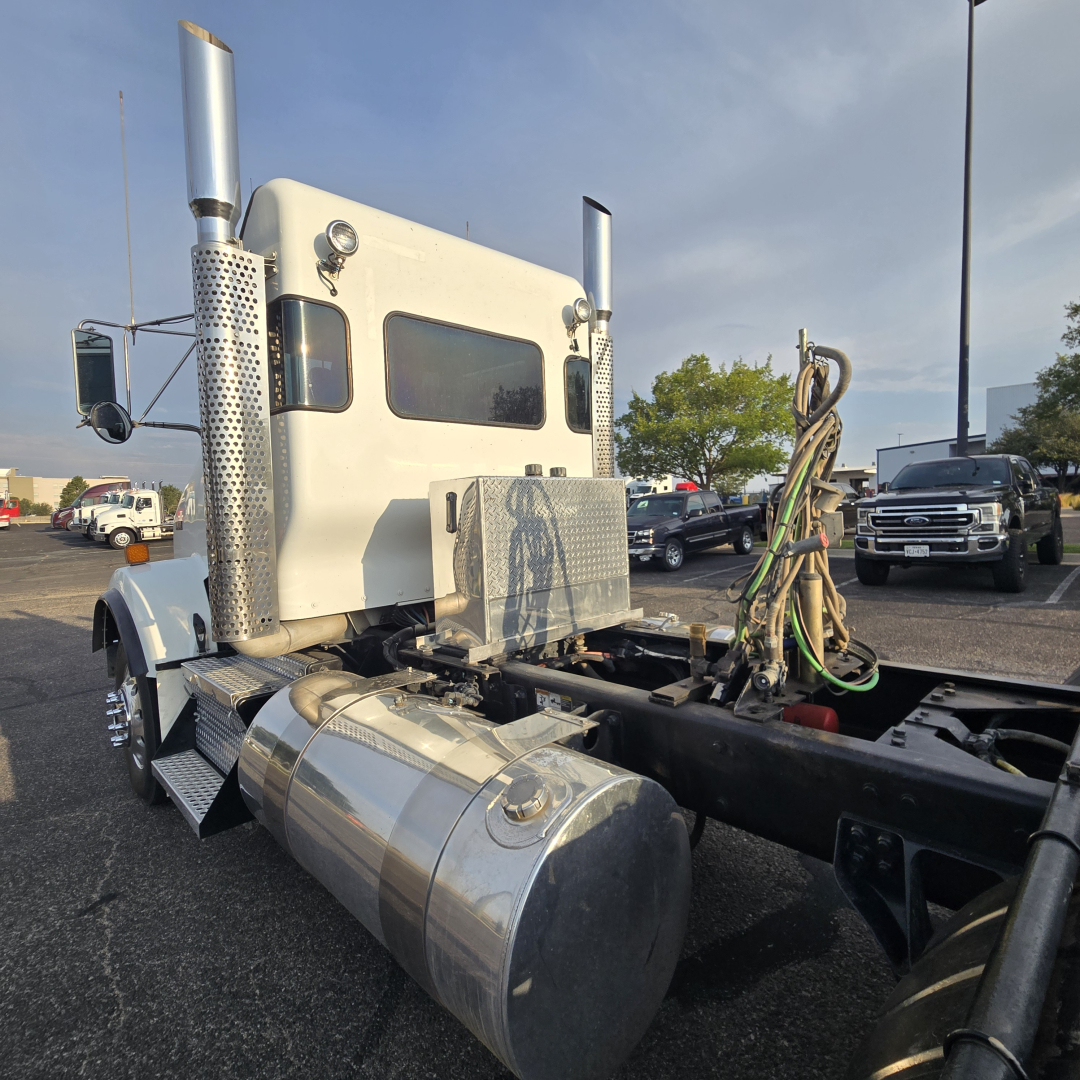 Kenworth T800 tractor cab side view showing chrome fuel tank, exhaust stacks, and wiring near rear chassis; front cab with two square windows and air intake.