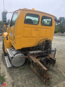 Cab and chassis truck rear view showing yellow Sterling Acterra cab with drivetrain and hitch, rusted implement attached to rear, hydraulic lines coiled, side steps