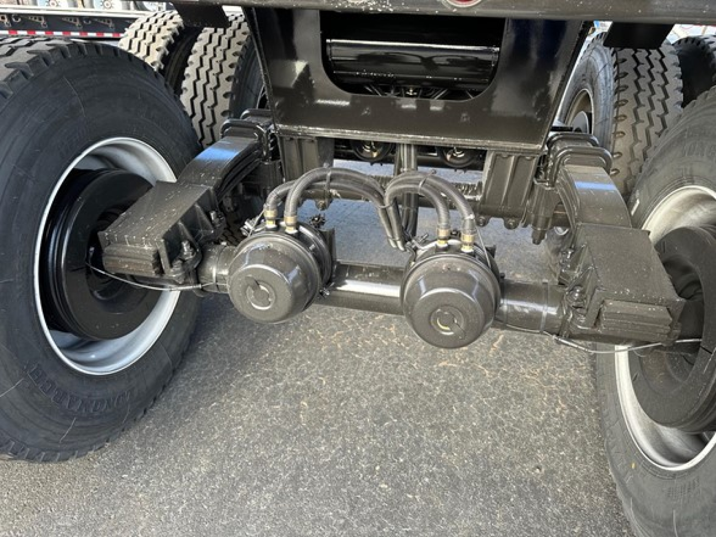 Underbody view of a dump trailer showing axles, suspension, and hoses with two circular components near center; tires on both sides.