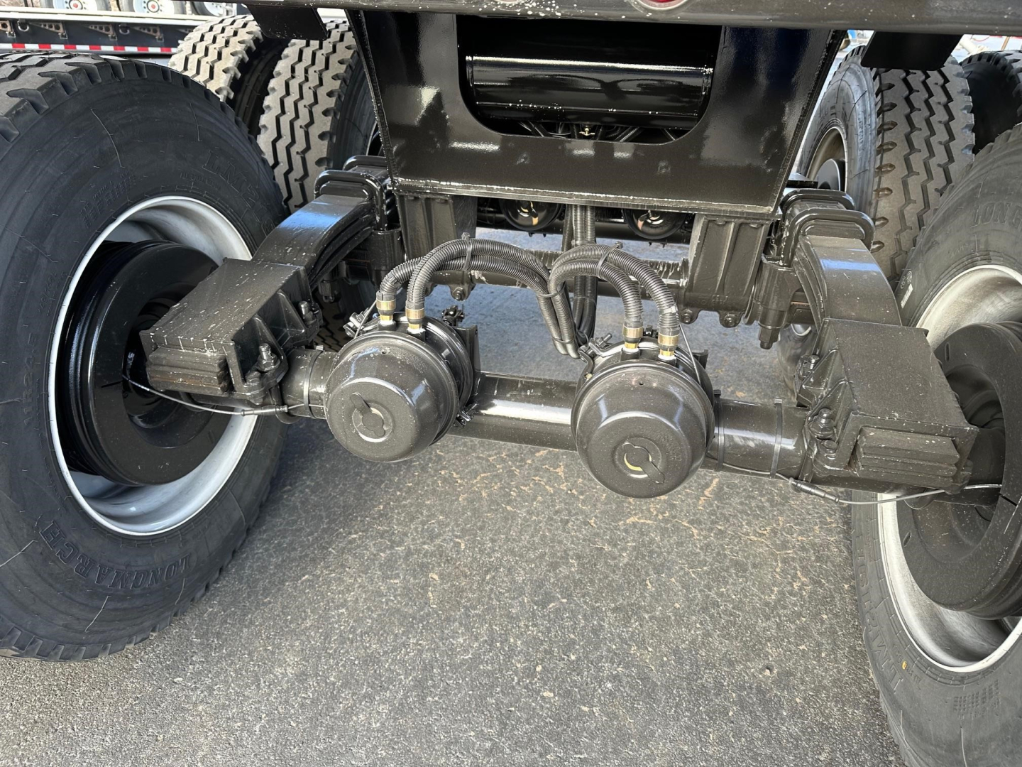 Undercarriage of a dump trailer showing a tandem axle with brake chambers and air lines, heavy-duty suspension, and tires. Visible components include two round brake chambers and several air hoses connected to a central manifold area.