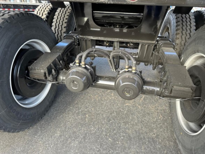 Underbody view of a dump trailer showing axles, suspension, and hoses with two circular components near center; tires on both sides.
