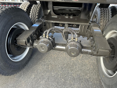 Undercarriage of a dump trailer showing a tandem axle with brake chambers and air lines, heavy-duty suspension, and tires. Visible components include two round brake chambers and several air hoses connected to a central manifold area.