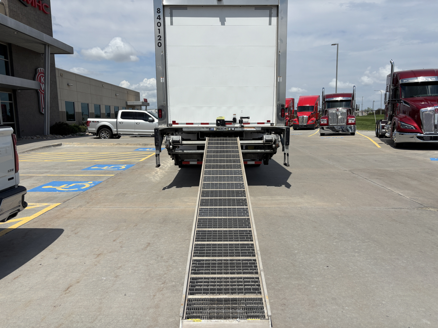 Rear view of a refrigerated Kenworth T380 with a long metal loading ramp extending to the ground, showing the back of the truck and surrounding parking area.
